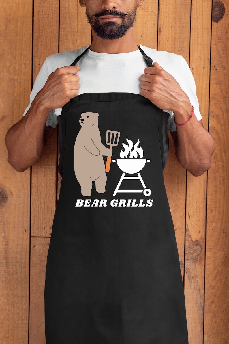Pu&ograve; includere: Grembiule nero con una grafica bianca di un orso che tiene una spatola e una griglia con fiamme. Il testo "BEAR GRILLS" &egrave; sotto la grafica.
