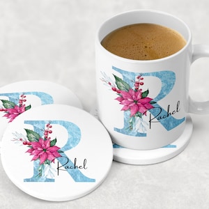 Pode incluir: Caneca de cerâmica branca com um design de aquarela azul e branco da letra R e uma flor de poinsettia. A caneca tem o nome Rachel escrito nela. A caneca está sobre um descanso de copo de cerâmica branca com o mesmo design.
