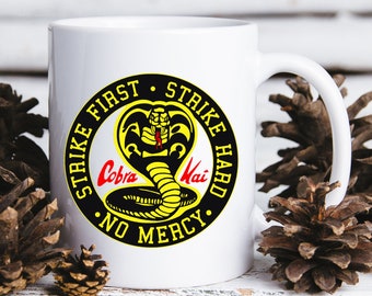 May include: White ceramic mug with a black and yellow Cobra Kai logo. The logo features a cobra with red eyes and the text "Strike First, Strike Hard, No Mercy".