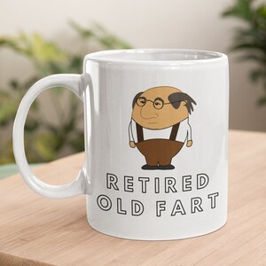 K&ouml;nnte beinhalten: Wei&szlig;e Keramiktasse mit einer Karikatur eines &auml;lteren Mannes mit Brille und Hosentr&auml;gern. Auf der Tasse steht in schwarzer Schrift "RETIRED OLD FART". Die Tasse steht auf einer Holzoberfl&auml;che.