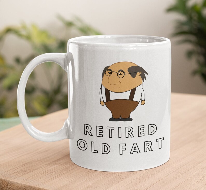K&ouml;nnte beinhalten: Wei&szlig;e Keramiktasse mit einer Karikatur eines &auml;lteren Mannes mit Brille und Hosentr&auml;gern. Auf der Tasse steht in schwarzer Schrift "RETIRED OLD FART". Die Tasse steht auf einer Holzoberfl&auml;che.
