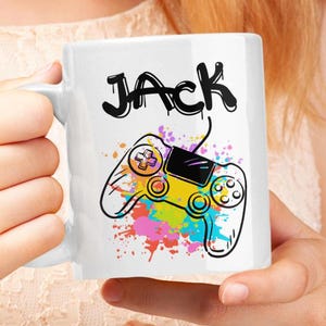 Puede incluir: Taza de cerámica blanca con un diseño de salpicaduras de pintura coloridas y un mando de videojuego en blanco y negro. El nombre "JACK" está escrito en letras de estilo graffiti negro.