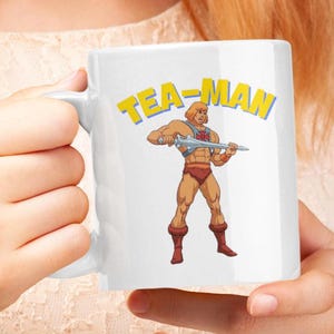 May include: White ceramic mug featuring a cartoon illustration of He-Man wielding a sword. The text "TEA-MAN" is printed in yellow above the image.