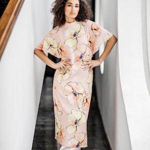 May include: A woman wearing a pink maxi dress with a floral print. The dress has a high neck and short sleeves. She is standing in a hallway with white walls.
