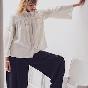 Op de afbeelding: Een persoon draagt een lichtgekleurde blouse met wijde mouwen en een zwarte broek met wijde pijpen. De blouse heeft een knoopsluiting aan de voorkant en een geplooide halslijn. Een parelketting en armband zijn zichtbaar. Zwarte enkellaarsjes met spitse neus maken de outfit compleet.
