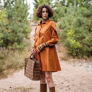 Puede incluir: Una persona con un vestido camisero naranja con mangas largas y puños a rayas. Lleva un bolso tote a juego con un patrón geométrico y botas marrones. La escena tiene lugar al aire libre.