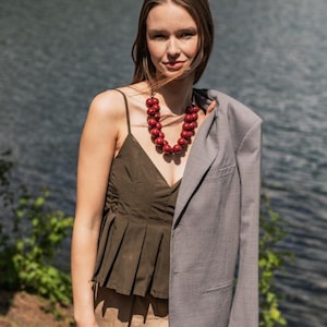 Op de afbeelding: Een donker olijfgroene geplooide camisole met dunne bandjes, een grijze blazer over één schouder gedrapeerd en een ketting met meerdere grote, glanzende, dieprode, bolvormige kralen in een meerstrengs ontwerp rond de hals.