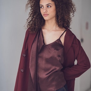 Op de afbeelding: Een bordeaux blazer wordt gedragen over een bijpassende satijnen top met spaghettibandjes. Het model draagt ook een donkerblauwe jeans. De outfit is een monochrome look met focus op textuur en kleur.