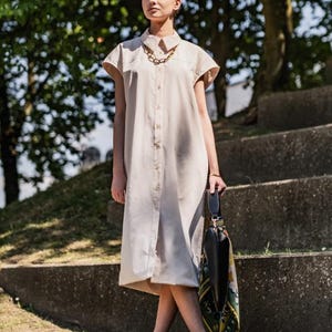 Op de afbeelding: Een beige midi-lengte overhemdjurk met korte mouwen, een klassieke kraag en een knoopsluiting over de gehele lengte. Het kledingstuk is gestyled met een grove goudkleurige ketting en een zwarte handtas met een bedrukte zijden sjaal eraan.