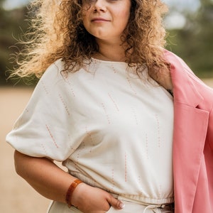May include: A woman with curly hair is wearing a white top with a pink pattern, white trousers, and a pink blazer. She is wearing a brown bracelet on her wrist.