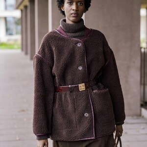 Op de afbeelding: Bruine fleecejas met bordeauxrode bies en bijpassende riem. De jas wordt gecombineerd met een bruine broek en een bruine coltrui. Een bruine handtas maakt de outfit compleet.