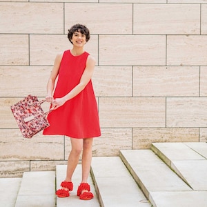 Peut inclure: Une femme portant une robe rouge sans manches et des sandales rouges tient un sac fourre-tout à motif floral devant un mur de carreaux beiges.