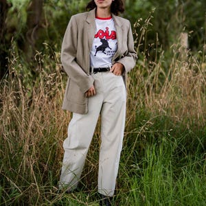 Op de afbeelding: Een persoon draagt een beige blazer over een wit t-shirt met een rode kraag en de tekst "Lois" en een stierafbeelding. Ze dragen ook lichtgekleurde broeken en een zwarte riem. De achtergrond is een veld met hoog gras.