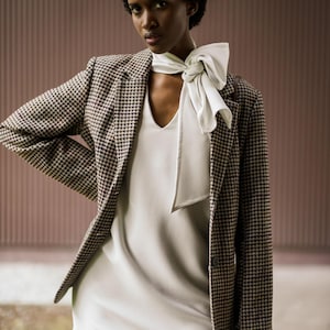 Op de afbeelding: Een vrouw draagt een blazer met pied-de-poule over een witte jurk. Een witte sjaal is om de nek geknoopt. De blazer heeft een klassiek patroon in bruin- en zwarttinten. De jurk heeft een V-hals.