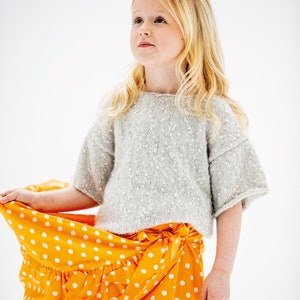 Op de afbeelding: Een jong meisje draagt een lichtgrijze trui met witte stippen en een feloranje rok met witte stippen en een gerimpelde zoom.