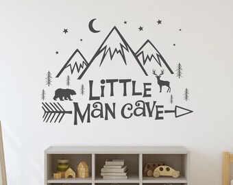 Op de afbeelding: Een grijze en witte muursticker met een bergketen, een halve maan, sterren, bomen, een beer, een hert, een pijl en de tekst "Little Man Cave".