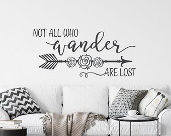 Puede incluir: Un sofá blanco con almohadas y una manta en una sala de estar con una calcomanía de pared que dice "Not all who wander are lost" con una flecha y detalles florales.