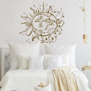Può includere: Un letto bianco con una coperta di uncinetto bianca e un cesto di vimini bianco. La parete dietro il letto ha un adesivo da parete sole e luna dorato con stelle.