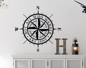 Puede incluir: Una calcomanía de pared negra con forma de rosa de los vientos con el texto "NOT ALL WHO WANDER ARE LOST" está montada en una pared blanca. La brújula está encima de un mueble blanco con cuatro puertas. Los elementos decorativos incluyen una letra "H" de metal, una linterna y maletas apiladas.