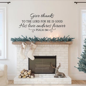 Puede incluir: Una chimenea de ladrillo blanco con una repisa de madera decorada con una guirnalda verde. Hay dos calcetines blancos colgando de la repisa. La chimenea tiene una pantalla de metal negro y una pila de leña delante. Una figurita de ciervo está sentada en el hogar. La pared sobre la chimenea tiene una calcomanía de vinilo negro con el texto "Give thanks to the Lord for he is good. His love endures forever. Psalm 136:1."