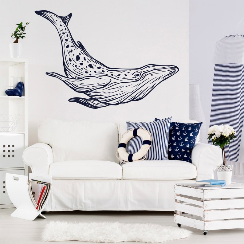 May include: A white couch with blue and white striped pillows and a life preserver pillow. The wall behind the couch has a black and white whale decal. The couch is in a living room with a white wooden side table and a white rug.
