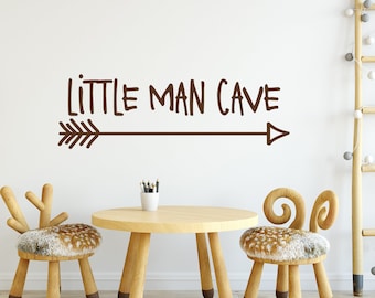 Op de afbeelding: Een bruine muursticker met de tekst "LITTLE MAN CAVE" en een pijl die naar rechts wijst. De sticker bevindt zich boven een houten tafel met twee houten stoelen met harige zittingen.