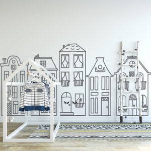 May include: A white wooden clothing rack with two striped shirts hanging on it. The rack is in front of a white wall with a black and white drawing of houses. A white wooden ladder with white round ornaments is on the right side of the image.