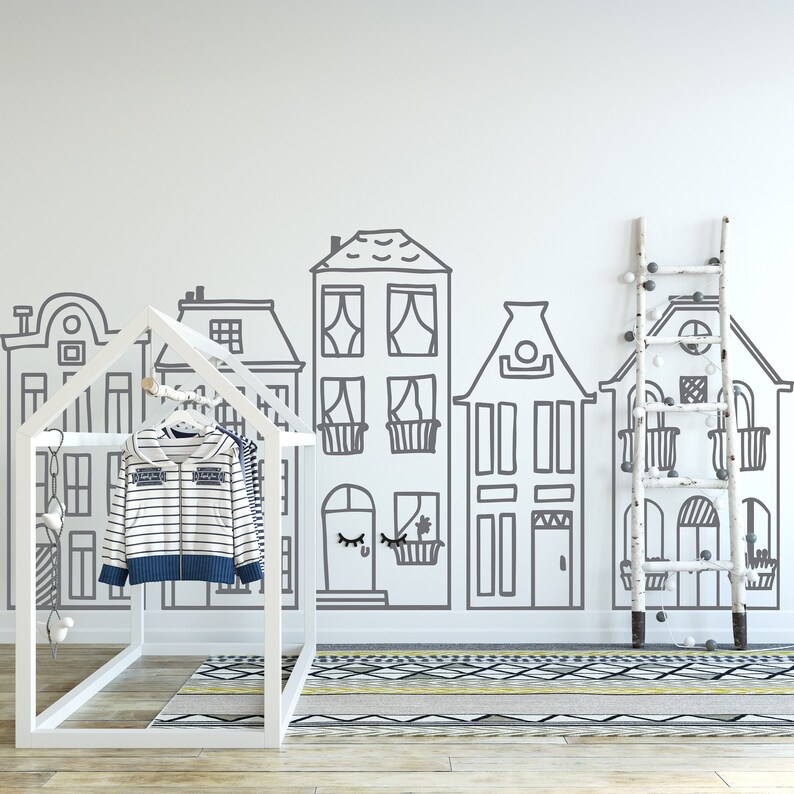 May include: A white wooden clothing rack with two striped shirts hanging on it. The rack is in front of a white wall with a black and white drawing of houses. A white wooden ladder with white round ornaments is on the right side of the image.