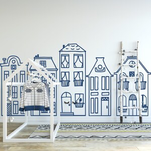 May include: A white wooden playhouse with a blue and white striped shirt hanging on a hanger inside. The playhouse is in front of a white wall with a blue hand-drawn cityscape design.