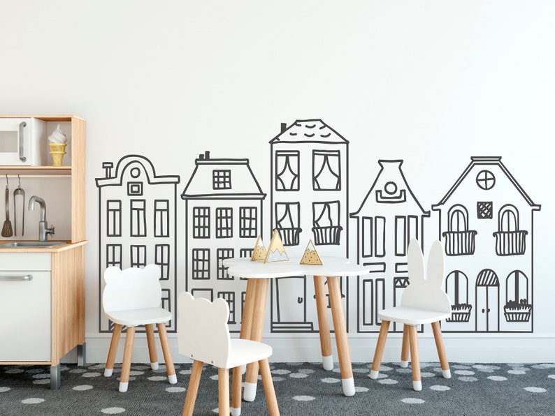 May include: A white playroom with a white wall featuring a black and white illustration of a row of houses. There are four white chairs with wooden legs and a small wooden table with a white top in the center of the room.