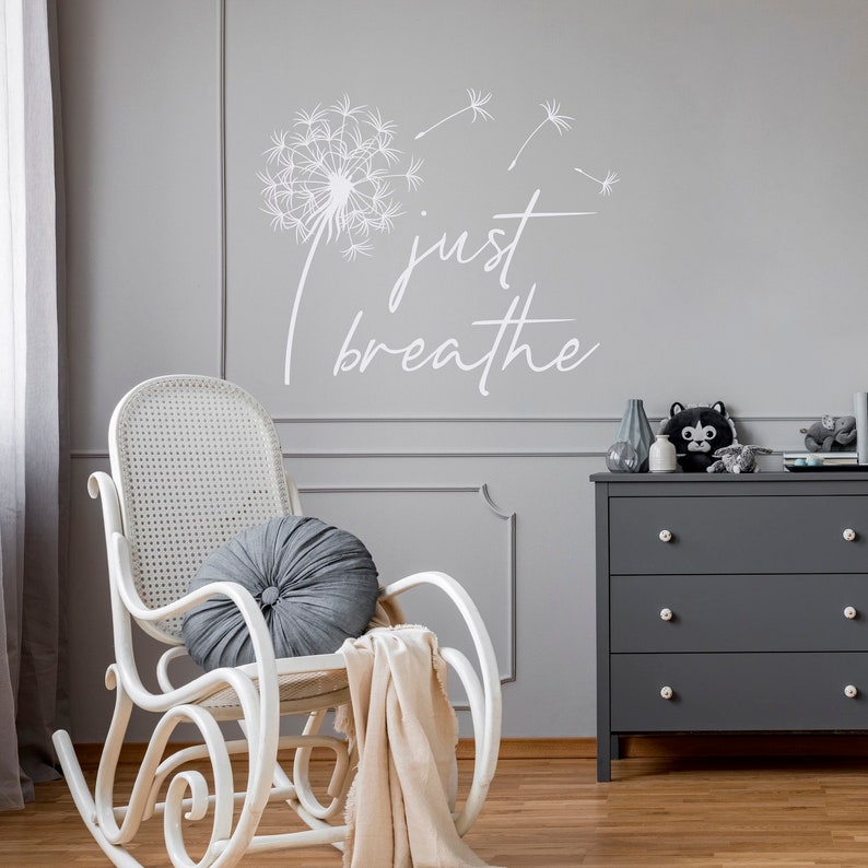 K&ouml;nnte beinhalten: Wei&szlig;er Wandaufkleber mit einem L&ouml;wenzahn und dem Text "just breathe". Im Vordergrund steht ein wei&szlig;er Schaukelstuhl mit einem grauen Kissen und einer beigefarbenen Decke.