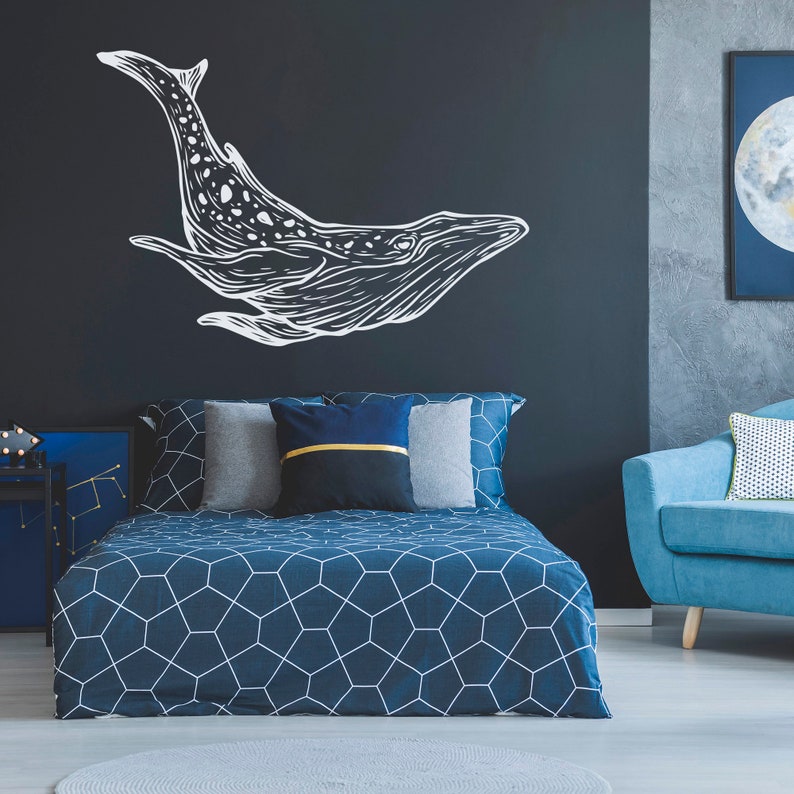 May include: A blue bed with a geometric patterned duvet cover and white pillows. The headboard is against a dark blue wall with a white whale silhouette decal. A blue armchair with a white pillow is in the corner of the room.