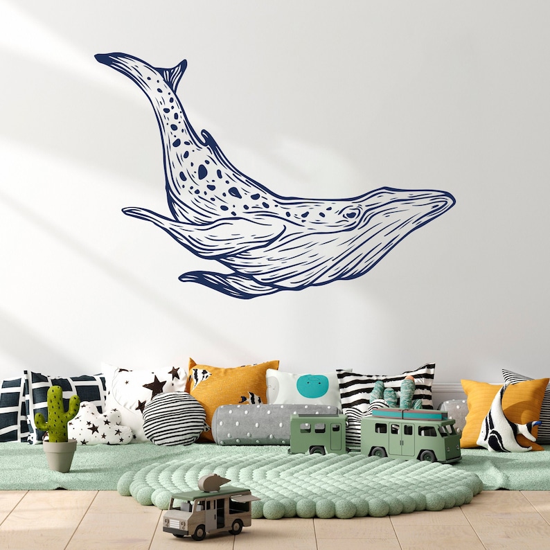 May include: A blue whale illustration wall decal, a popular choice for children's rooms, is displayed on a white wall above a play area with pillows and toy trucks.