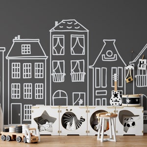 May include: A black and white illustration of a city street with houses drawn on a dark gray wall. A white storage unit with three compartments is in front of the wall. The compartments contain toys, including a toy truck, a toy drum, and a toy guitar.