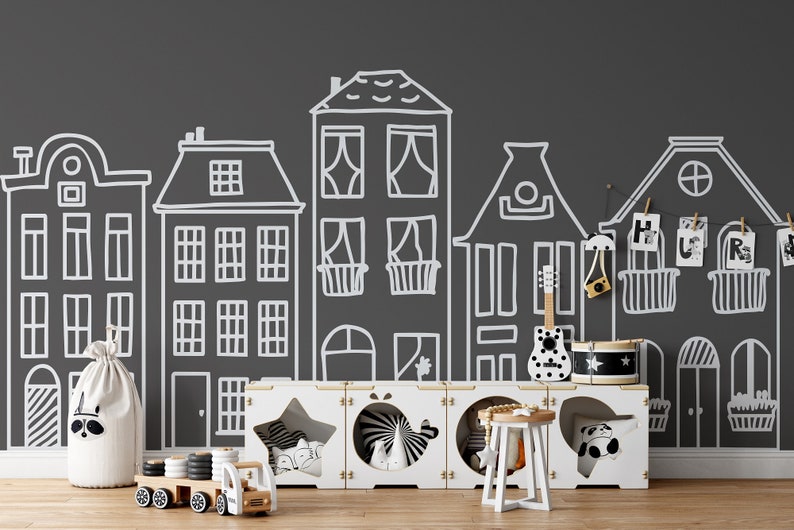 May include: A black and white illustration of a city street with houses drawn on a dark gray wall. A white storage unit with three compartments is in front of the wall. The compartments contain toys, including a toy truck, a toy drum, and a toy guitar.