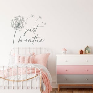 K&ouml;nnte beinhalten: Ein wei&szlig;es Metallbettgestell mit einer rosa Decke und Kissen. Die Wand hinter dem Bett hat einen grauen L&ouml;wenzahn und den Text "just breathe" in Grau.