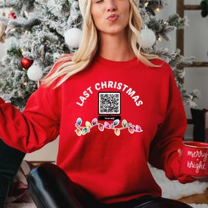 Op de afbeelding: Rood kersttrui met de tekst "Last Christmas" en een QR-code met de tekst "Scan me!" eronder. De trui heeft ook een string met gekleurde lampjes.