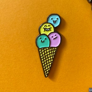 Può includere: Una chiave in stile vintage con un design elaborato e una finitura marrone, accanto a uno spillo in smalto giallo, rosa e blu di un cono gelato con un viso sorridente su ogni pallina.