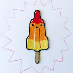 Può includere: Un ghiacciolo rosso, arancione e giallo con un viso sorridente ricamato su uno sfondo bianco.