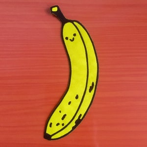 May include: A yellow banana-shaped patch with a smiling face and black spots. The patch has a black outline and a brown stem. The patch is likely for clothing or accessories.