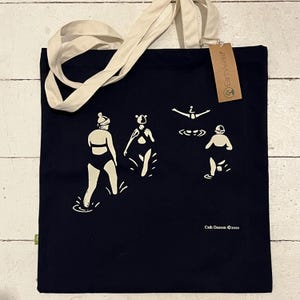 May include: A navy blue tote bag featuring a white design of people swimming in the sea. The bag has a white cotton handle and a small brown tag that says "Earth Aware". The bottom of the bag has the text "Cath Deeson ©2020".