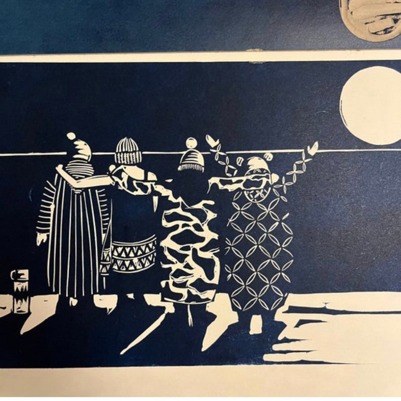 May include: A linocut print in shades of navy blue and white depicts a group of figures with arms around each other. The figures wear patterned clothing and hats, standing beneath a white moon. A mug is visible on the left.