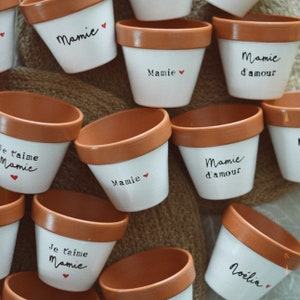 Op de afbeelding: Een verzameling kleine, witte keramische bloempotten met een terracotta rand. Elke pot heeft een handgeschreven boodschap in zwarte inkt, zoals "Mamie" of "Je t'aime Mamie", met een klein rood hart. De potten zijn gerangschikt op een getextureerd, neutraal gekleurd oppervlak.