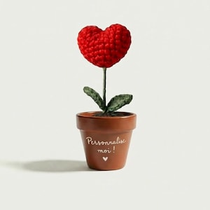 Può includere: Un fiore a forma di cuore all'uncinetto fatto a mano in un piccolo vaso marrone. Il fiore rosso a forma di cuore è posto su uno stelo verde con foglie verdi. Il vaso ha le parole "Personnalise moi !" e un simbolo di cuore.