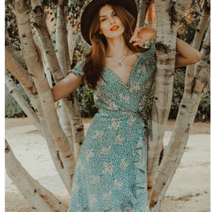 May include: A teal wrap dress with a floral pattern and ruffled trim. The dress has short sleeves and a V-neckline. A brown hat and necklace complete the look. The dress is tied at the waist.