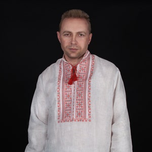 May include: A long-sleeved, off-white linen shirt featuring a stand-up collar and a central vertical panel decorated with intricate red and black geometric embroidery. The shirt includes a red drawstring with tassels at the neck and matching embroidery on the cuffs.