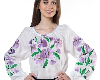 Puede incluir: Una blusa blanca de manga larga con bordados florales morados y hojas verdes en el pecho y las mangas. El escote incluye un cordón con borlas moradas. La blusa se combina con una falda negra lisa.