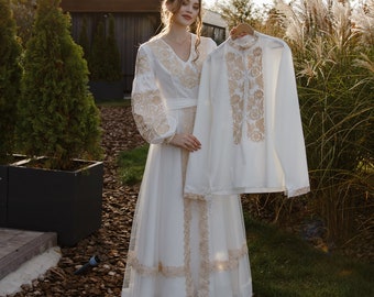May include: A woman in a white embroidered dress holds a white embroidered shirt with gold embroidery. The dress and shirt are traditional Ukrainian clothing.