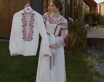 Könnte beinhalten: Ein weißes besticktes Hemd und ein weißes Kleid mit roter Blumenstickerei. Das langärmlige Hemd wird an einem Kleiderbügel gehalten. Das Kleid hat einen langen Rock und transparente Ärmel. Die Stickerei ist tiefrot.