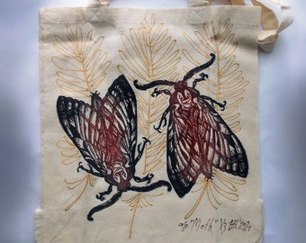 Op de afbeelding: Een witte tote bag met een zwarte en rode print van twee motten op een gouden achtergrond. De motten hebben ingewikkelde vleugelpatronen en zijn omringd door gestileerde varenbladeren. De tas heeft een datum en handtekening in zwarte inkt in de rechterbenedenhoek: "9/6/11/014" "X3 2021".